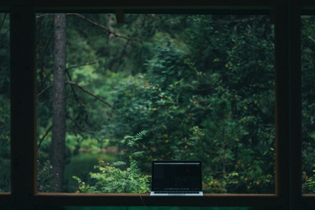 A view from a window showcasing lush green trees and foliage, with a laptop placed on a wooden table in the foreground.
